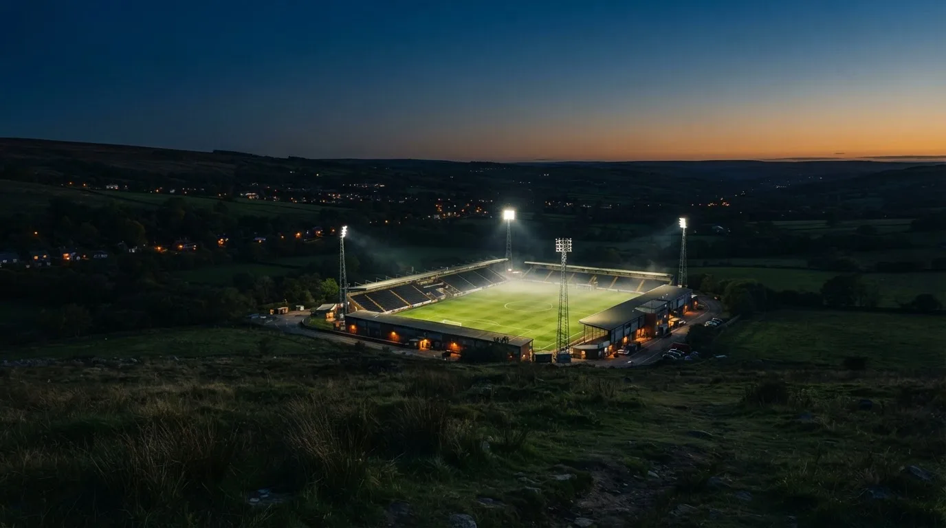 Ein Stadion unter Flutlicht bei Nacht, von einem Hügel aus der Ferne betrachtet, mit Dunst über dem Spielfeld.