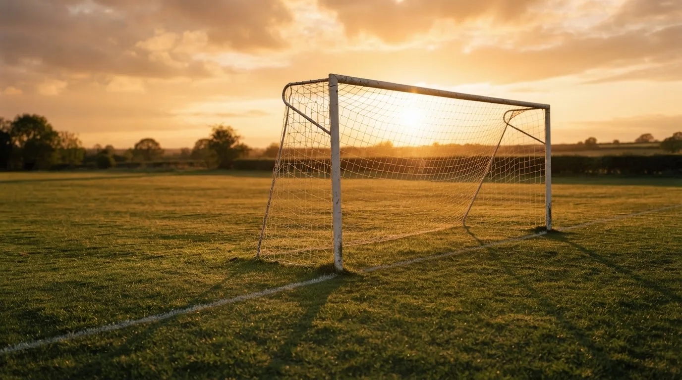 Ein Fußballtor mit Netz bei Sonnenuntergang auf einem leeren Platz, die Schatten der Torpfosten liegen lang auf dem Gras.