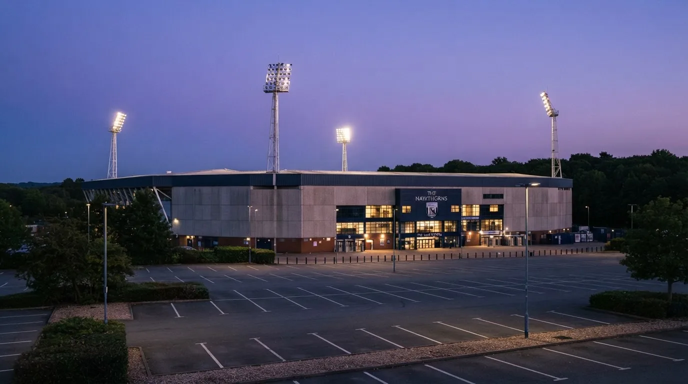 Ein Fußballstadion von außen in der Dämmerung, hohe Flutlichtmasten vor einem violetten Himmel, davor ein leerer Parkplatz.