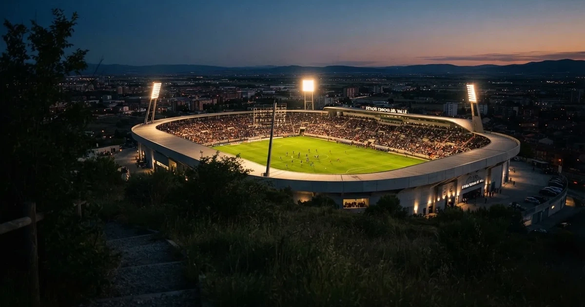 Ein Fußballstadion unter Flutlicht mit hellen Rängen, von einem Hügel aus gesehen, darüber ein dunkelblauer Abendhimmel.