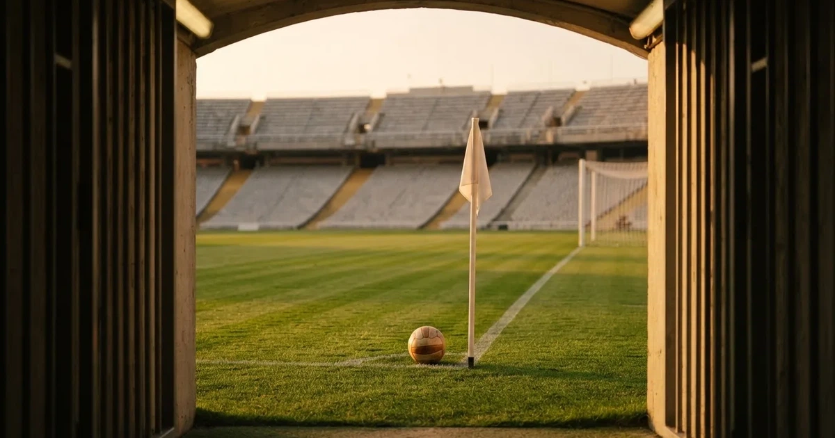 Blick durch ein Stadiontor auf einen grünen Rasen im Abendlicht, ein Ball ruht an der Eckfahne, die Tribünen sind leer.