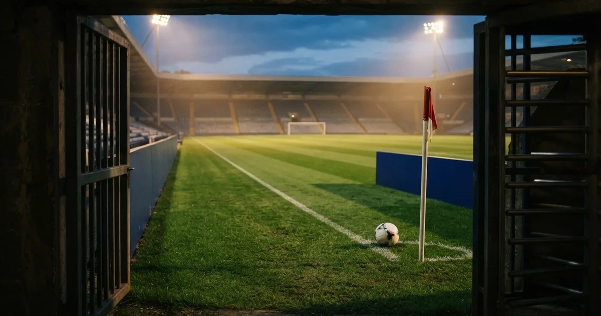 Blick durch ein Stadiontor auf einen grünen Rasen im Abendlicht, ein Ball liegt an der Eckfahne, die Ränge sind leer.