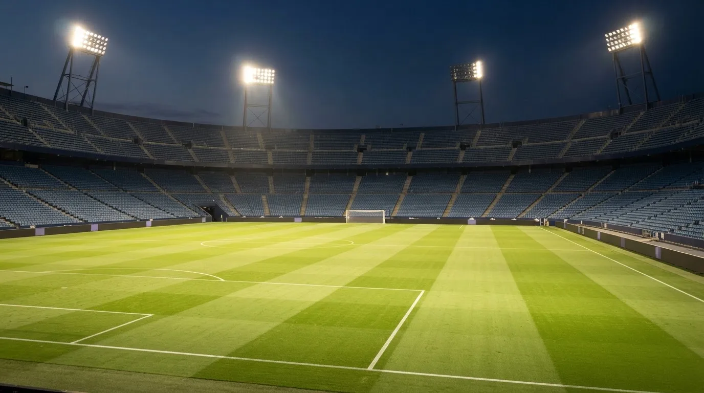 Eine leere Stadiontribüne unter Flutlicht aus der Ferne betrachtet, die Rasenfläche leuchtet grün in der Dämmerung.