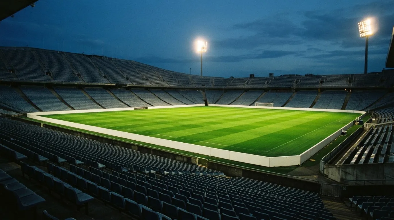 Eine leere Stadiontribüne unter Flutlicht aus der Ferne betrachtet, die Rasenfläche leuchtet grün in der Dämmerung.