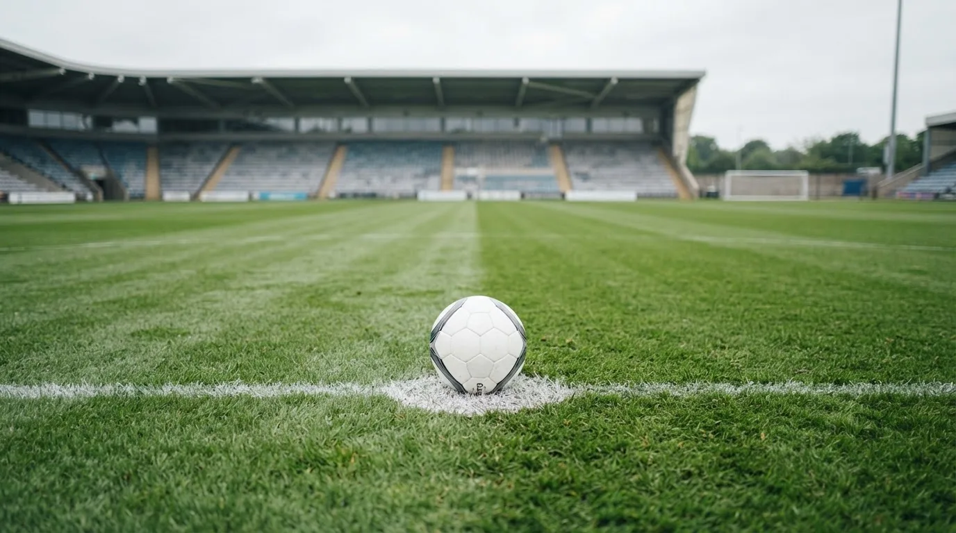 Ein Fußball liegt auf dem Anstoßpunkt eines leeren Rasenplatzes, im Hintergrund eine ruhige Tribüne unter bedecktem Himmel.