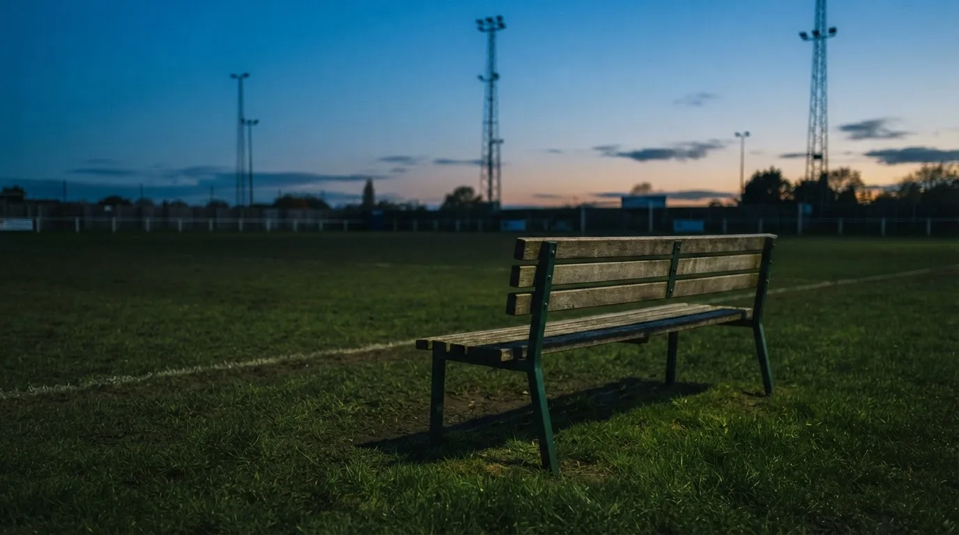 Eine leere Parkbank am Rand eines Fußballfelds in der Abenddämmerung, im Hintergrund ragen dunkle Flutlichtmasten in den Himmel.